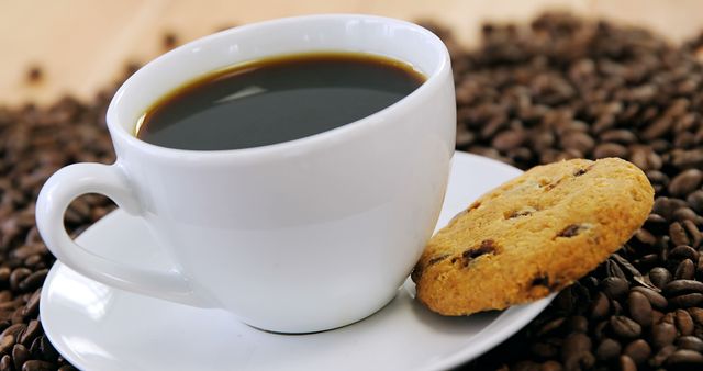 A cup of black coffee sits on a saucer surrounded by coffee beans, accompanied by a chocolate chip cookie. The arrangement invites a moment of relaxation or a quick coffee break with a sweet snack.
