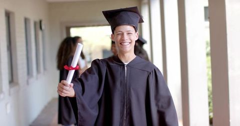 Smiling student wearing cap and gown holding diploma while walking campus corridor