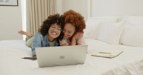 Laughing friends sharing laptop on cozy bed, studying and browsing together
