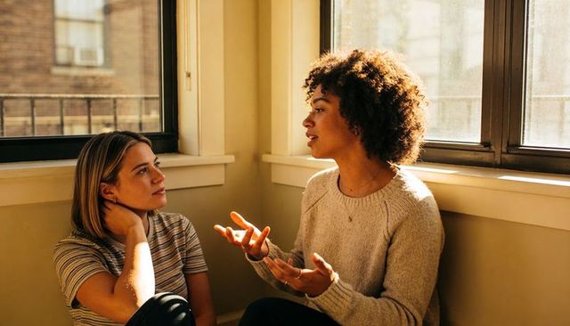 Two friends having heart-to-heart conversation in sunlit apartment corner
