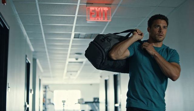 Fit man carrying gym duffel walking down corridor toward exit sign, fitness center interior