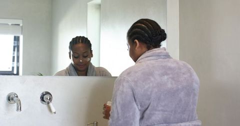 African american woman applying moisturizer at bathroom mirror for morning skincare routine