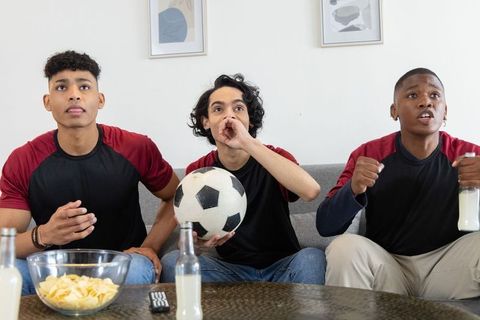 Diverse teens watching intense soccer match on couch, cheering and holding soccer ball