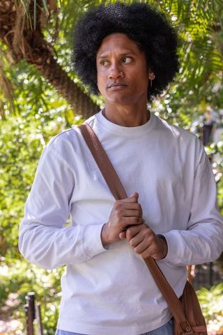 Young african american man holding bag strap standing under sunlit trees in park
