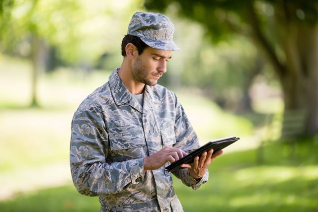 Soldier in camouflage uniform using digital tablet in a park on a sunny day. Ideal for use in articles about military technology, outdoor activities, modern communication in the armed forces, or professional use of digital devices. Can also be used in promotional materials for military recruitment or technology integration in the military.