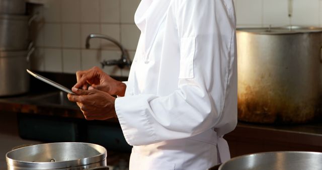 Chef wearing white uniform using digital tablet in a professional kitchen. Ideal for content on modern cooking technology, culinary arts, restaurant management, and professional chef workflows. Could be used for websites, blogs, and advertisements related to food industry advancements and kitchen technology.