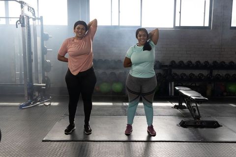 Two african american plus-size women stretching together in gym with weights and camaraderie