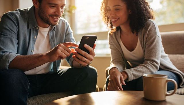 Couple sharing smartphone and smiling on cozy sofa during sunny morning coffee