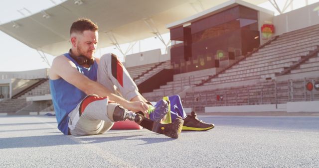 Caucasian disabled male athlete with prosthetic leg sitting and putting on sports shoes. professional runner training at sports stadium.
