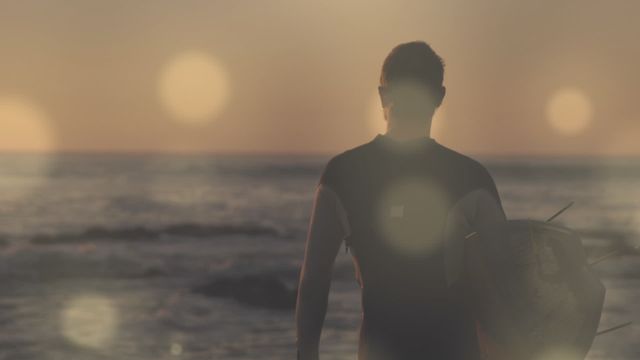 Surfer standing on beach holding board, looking at ocean during sunset. Perfect for content related to surfing, beach lifestyles, outdoor adventure promotions, and travel advertisements.