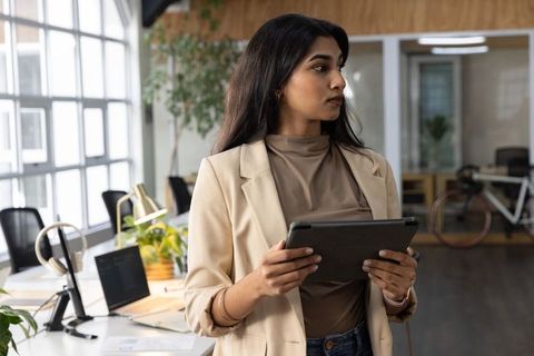 Confident indian businesswoman holding tablet in modern open-plan office with plants