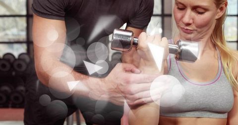 Woman lifting chrome dumbbell, trainer spotting and correcting form during gym session