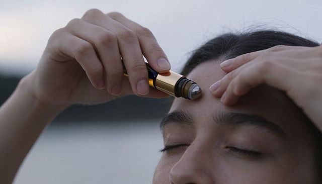 Applying amber roll-on serum to forehead outdoors by water, close-up wellness ritual