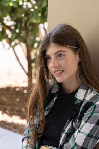 Young woman leaning on wall wearing plaid shirt and belt buckle in sunlit courtyard