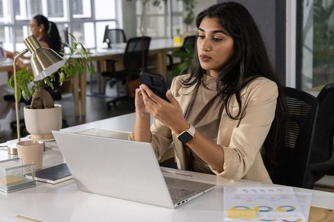 Indian professional checking smartphone while reviewing laptop charts in coworking office