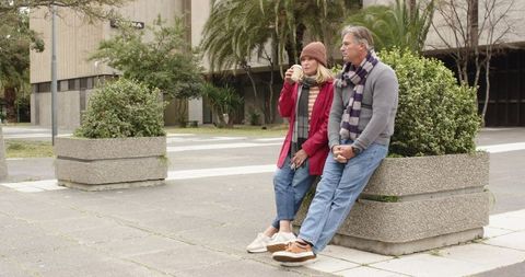 Senior couple sitting on urban planter sipping takeaway coffee wearing scarves and beanie