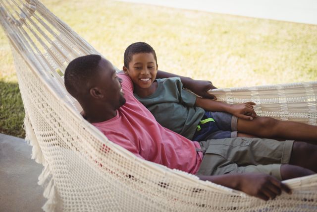 Father and son enjoying a relaxing moment on a hammock in a garden. Perfect for themes related to family bonding, outdoor leisure, summer activities, and parenting. Ideal for use in advertisements, blogs, and articles focused on family life, happiness, and togetherness.