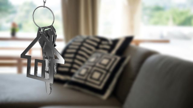 Visual capturing house keys with a key fob shaped as a house, hovering near a cozy, modern living room couch with stylish cushions. Useful for real estate agencies, home decor websites, property investment promotions, and depicting new homeownership.