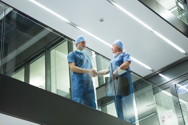 Two male surgeons in surgical attire are shaking hands in a hospital corridor, symbolizing teamwork and collaboration in a medical setting. This image can be used for promoting healthcare services, medical teamwork, professional collaboration, and hospital environments.