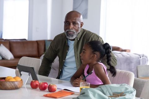 Older man helping young girl with homework at bright kitchen table using tablet, notebook