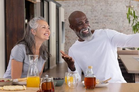 Mature diverse couple sharing sunny pancake breakfast at long wooden table in modern loft