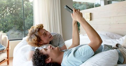 Happy Diverse Gay Couple Relaxing with Smartphone on Bed