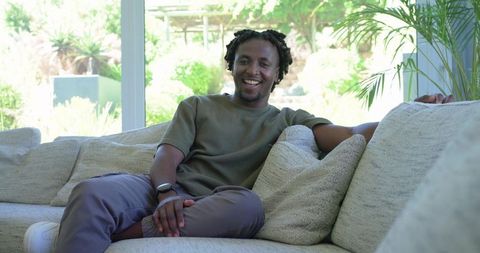 Relaxed african man smiling on modern sofa by window wearing smartwatch, casual lifestyle