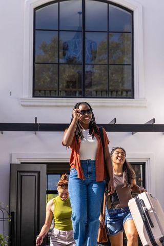 Friends walking down steps of modern house with luggage, african woman adjusting sunglasses