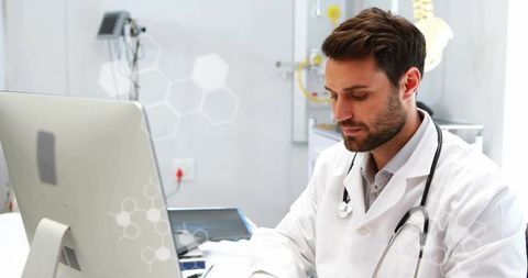 Focused male physician reviewing patient records on computer and tablet at desk in modern clinic