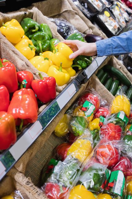 Hand selecting a fresh bell pepper from a grocery store shelf. Ideal for use in articles about healthy eating, grocery shopping tips, organic produce, and nutrition. Can also be used in marketing materials for supermarkets, food blogs, and health-related content.