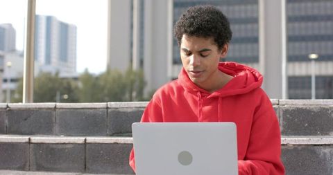 African american man in red hoodie working on laptop on campus steps in urban daylight