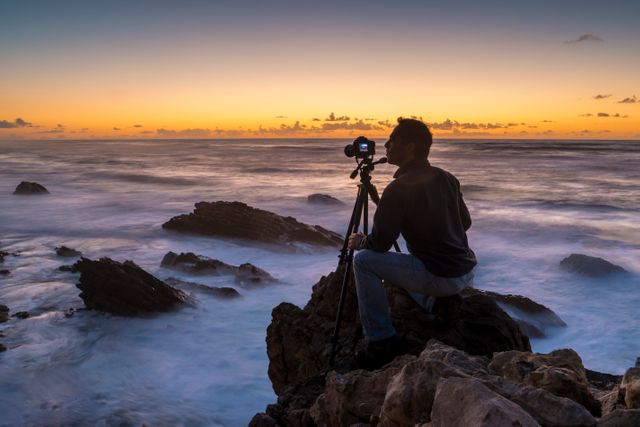 Photographer capturing stunning sunset at rocky seashore with a tripod. Ideal for travel, nature, photography, ocean and evening themes.