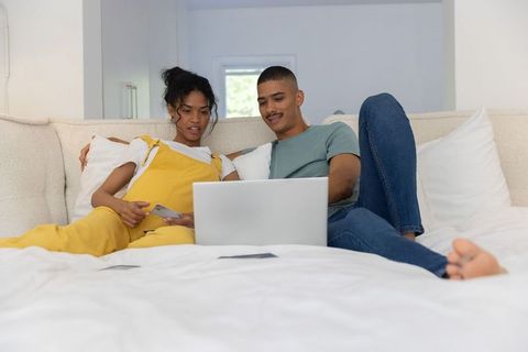 Young couple browsing laptop from bed holding credit card, cozy home shopping and leisure