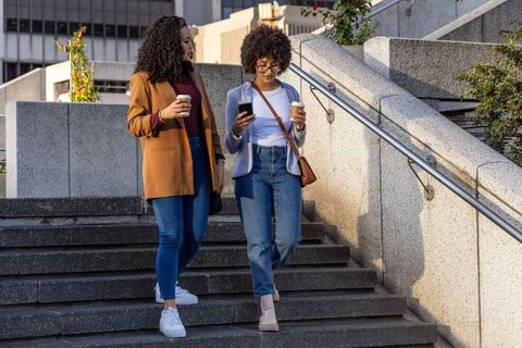 Friends walking down sunlit urban concrete stairs holding coffee and checking smartphone