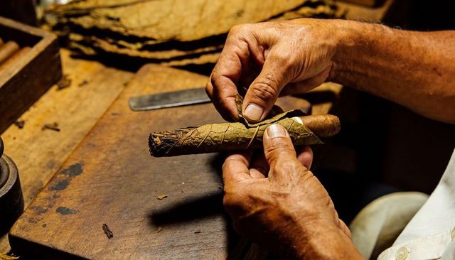 Artisan hands rolling cigar with tobacco leaf on rustic wood bench closeup