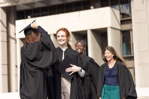 Female graduates celebrating on campus plaza wearing caps and gowns holding diplomas