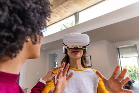 African american woman guiding friend wearing vr headset in bright modern living room