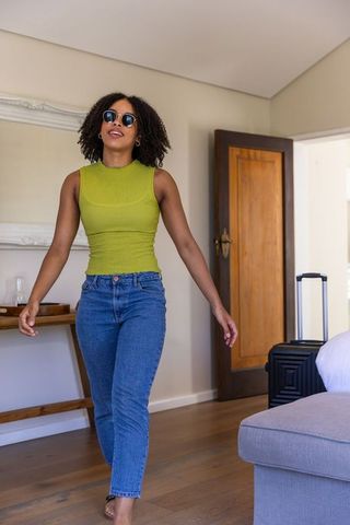 African american woman walking into sunlit hotel room wearing sunglasses with suitcase