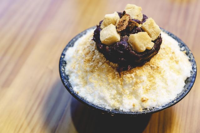 The image shows a bowl of traditional Korean Patbingsu, featuring a generous serving of shaved ice topped with sweet red beans, mochi, and a sprinkling of nuts or flakes. This popular Korean dessert is known for being both refreshing and satisfying, especially during the summer months. The image can be used for food blogs, recipe websites, cultural articles, and advertisements promoting Asian desserts or summer treats.