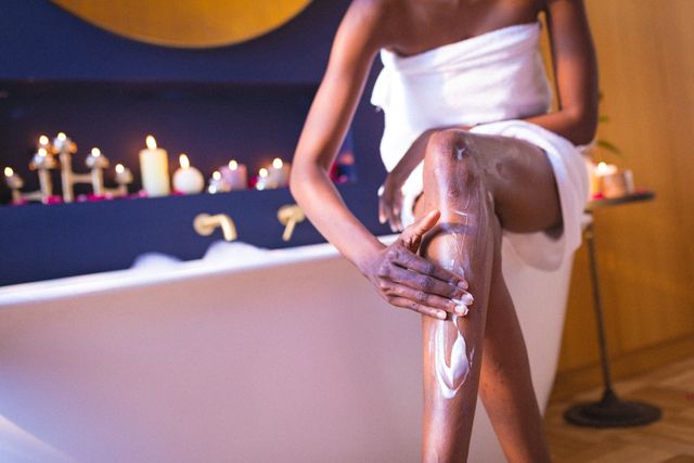 Midsection of african american young woman wrapped in towel applying cream on legs in bathroom. unaltered, people, lifestyle, home and self care concept.