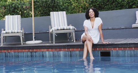 Smiling african american woman relaxing at pool edge dipping feet in blue water