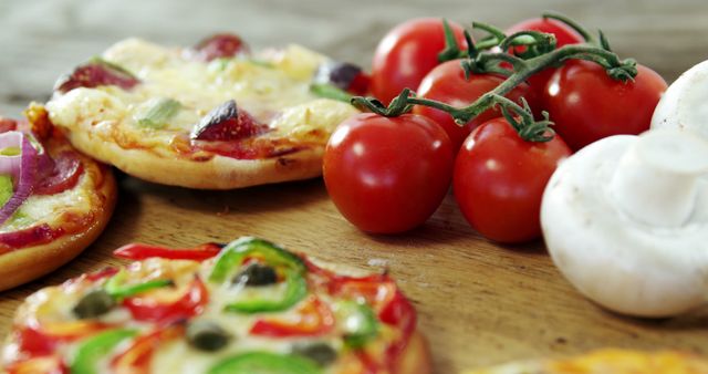 Mini pizzas with various toppings lie on a wooden table next to fresh tomatoes and mushrooms. This image is ideal for food blogs, restaurant menus, recipe books, or advertisements promoting fresh ingredients and Italian cuisine.