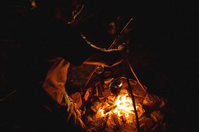 Young male survivalist cooking meal over burning campfire in forest at night. Ideal for content related to outdoor adventures, survival skills, camping, and nature activities. Can be used in articles, blogs, or advertisements focusing on wilderness experiences and survival techniques.