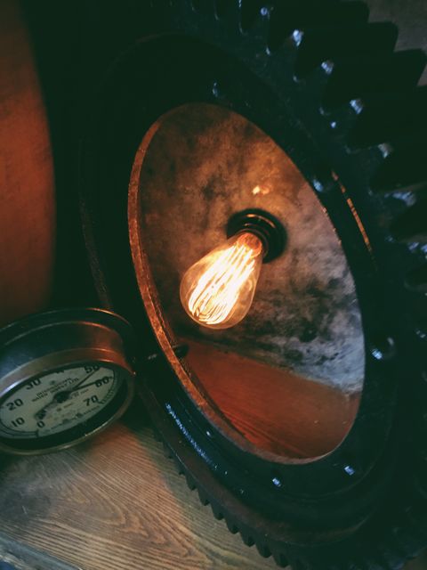 This image depicts a vintage-style light bulb and a machine gauge placed against a worn wooden background, creating an industrial aesthetic. The warm glow of the light bulb complements the rustic elements and mechanical parts. Ideal for use in articles, blogs, and promotional material related to interior design, retro-futurism, or steampunk themes.