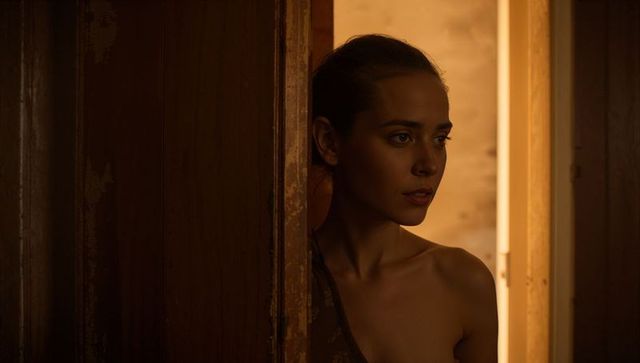 Young woman peering through weathered doorframe in warm amber low light, intimate mood