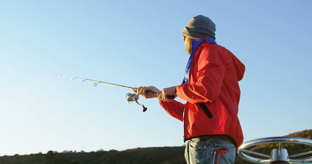 A man in a red jacket and beanie fishing with a rod from a boat during sunset. Scenic mountain in background. Ideal for advertisements related to outdoor activities, recreational fishing, travel, mindfulness, and nature exploration.
