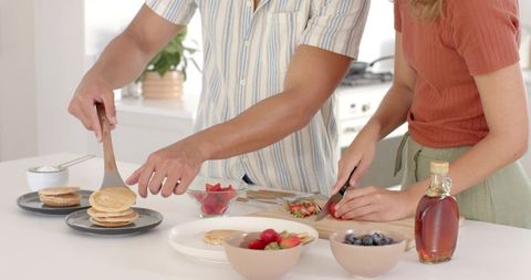 Diverse couple preparing pancakes and fruits in modern kitchen setting
