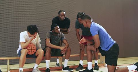 Young diverse male basketball teammates rallying around kneeling captain in gym huddle
