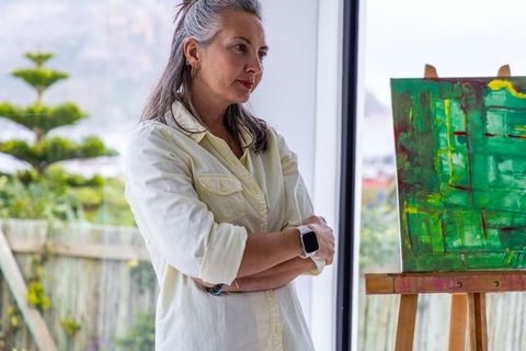 Mature woman contemplating abstract green painting by sunlit window wearing smartwatch