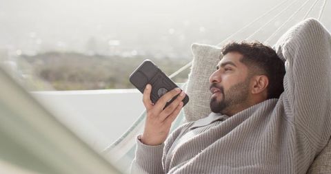 Indian man reclining in hammock on terrace using smartphone for voice message